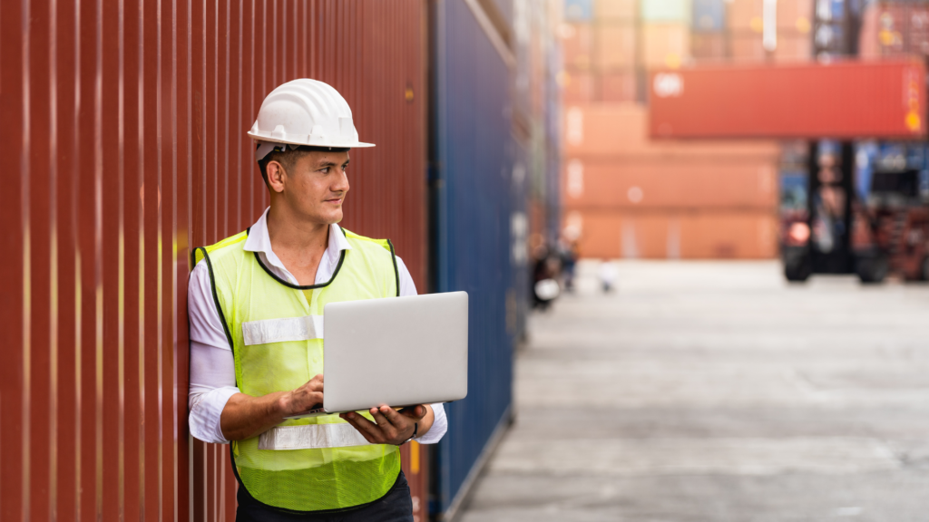 Logistics worker in safety vest and helmet using a laptop at a shipping container yard